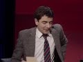 One of the most loved clips from Rowan's vast back catalogue, a hilarious sketch where the angry teacher played by Rowan invites a father in to talk about the trouble with son... Whether mesmerising us with the sheer visual mastery of Mr. Bean, beguiling us with the acerbic wit of Edmund Blackadder, or simply entertaining us as the suave, but rather hapless British Secret Agent Johnny English, you surely won't have escaped the comic genius that is Rowan Atkinson. In Rowan Atkinson Live, co-written with Richard Curtis (4 Weddings & a Funeral, Notting Hill, Love Actually) and Ben Elton, Atkinson runs the whole gamut of his remarkably versatile 30 year career, with sketches, mimes and monologue's that are guaranteed to have you shedding tears of laughter. Performing live on stage alongside "straight man" Angus Deayton, the show features a number of original and familiar routines, including sketches that appeared in the original Mr. Bean series.