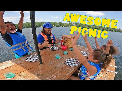 Group of kids in life jackets cheer around a picnic table on a lake, with checkered plates and a wicker basket nearby.