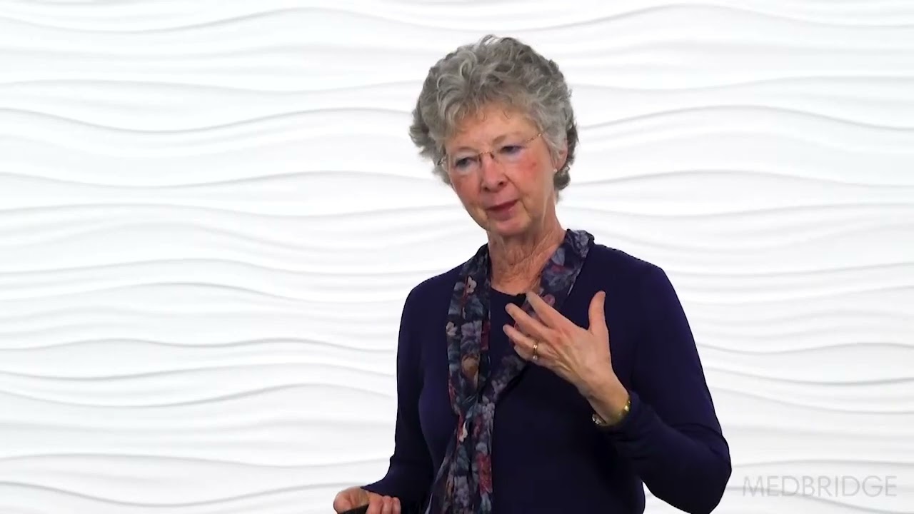 An older woman with gray curly hair and glasses gestures near her chest in front of a white wall, suitable for medbridge healthcare education.