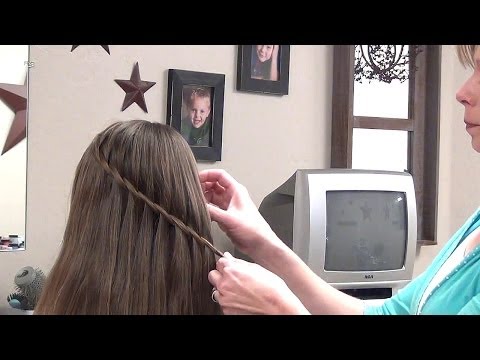 Person demonstrating a twist-waterfall hairstyle on a child's long hair in a home setting, with framed photos and a television in the background, highlighting quick and easy hairstyle techniques for busy parents.