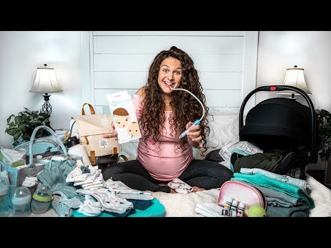 Pregnant woman sitting cross‑legged on a bed, smiling and using a breast pump while surrounded by baby items and a diaper bag in a cluttered nursery setup.