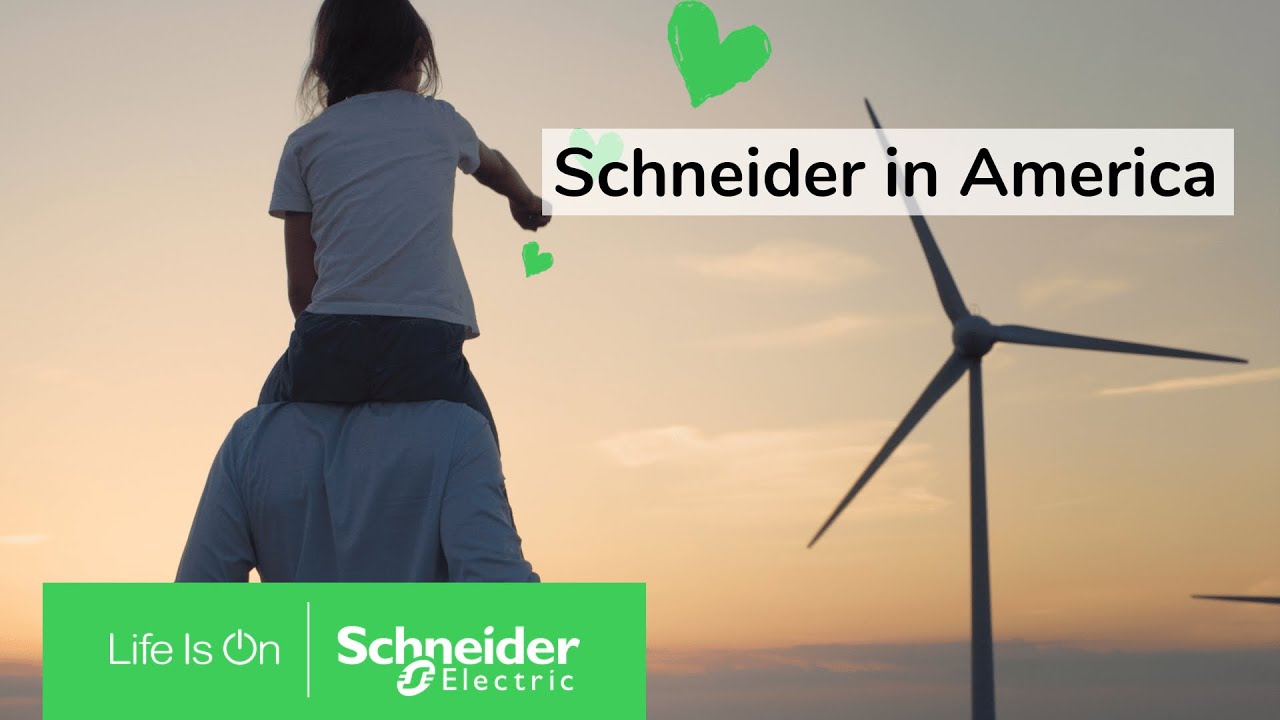 Silhouette of a man carrying a child on his shoulders who’s pointing toward a large wind turbine as the sun is setting in the background.