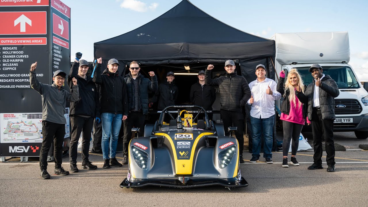 a group of happy people stand around a racing car with hands and thumbs raised in celebration