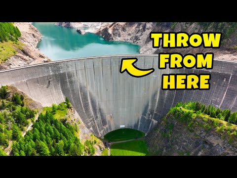 A massive curved concrete dam with turquoise water behind it and a large yellow sign reading 'THROW FROM HERE' on the upper right side.