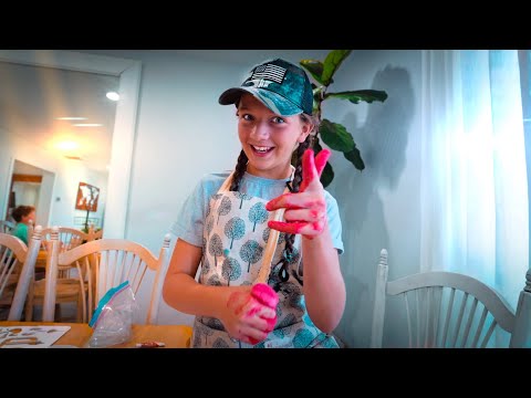 Girl with braids and a cap smiles at the camera while painting, her hands covered in bright pink paint, in a cozy dining room workspace.