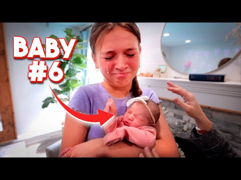 Mother cradles a newborn baby wearing pink in a cozy room, with bold 'BABY #6' text overlay and a hand reaching toward the baby.