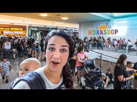 Woman wearing a baby carrier takes a selfie in a crowded mall near Build-A-Bear Workshop sign.