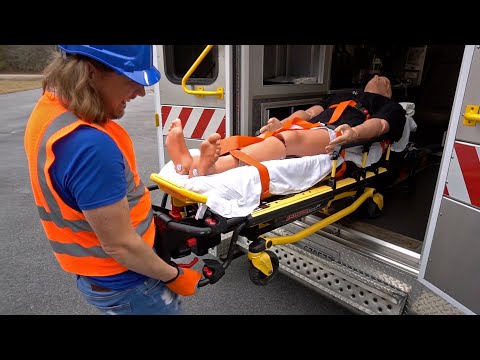 Emergency responder in an orange safety vest and blue helmet wheels a patient on a stretcher toward an ambulance entrance.