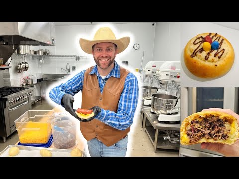 Smiling man in a cowboy hat and brown vest builds a sandwich in a commercial bakery kitchen, with mixers and cookware in the background.
