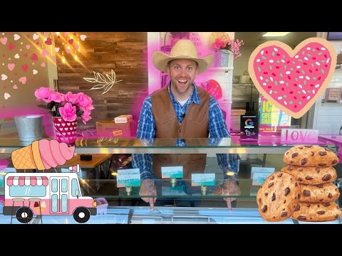 Smiling man in a cowboy hat and brown vest behind a pink, heart-themed ice cream shop counter, with cookies and decorations nearby.