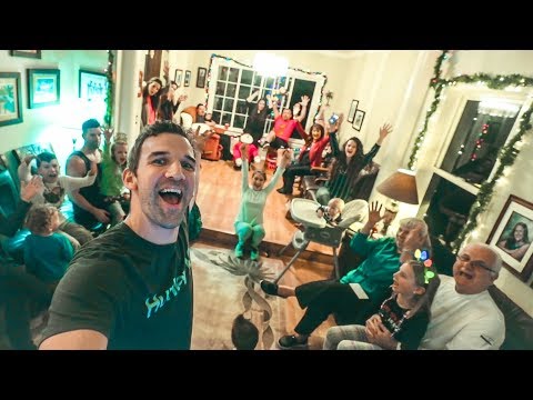 Man in foreground takes a selfie at a festive family Christmas gathering; everyone cheers with raised hands in the living room.