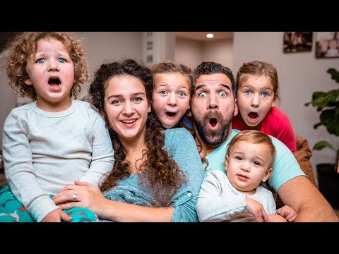 A smiling family of six sits close together on a couch, all with surprised, wide-eyed expressions and open mouths.