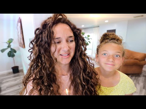 Mother with long curly hair and daughter with a bun smiling in a bright living room, indoors in a home setting.