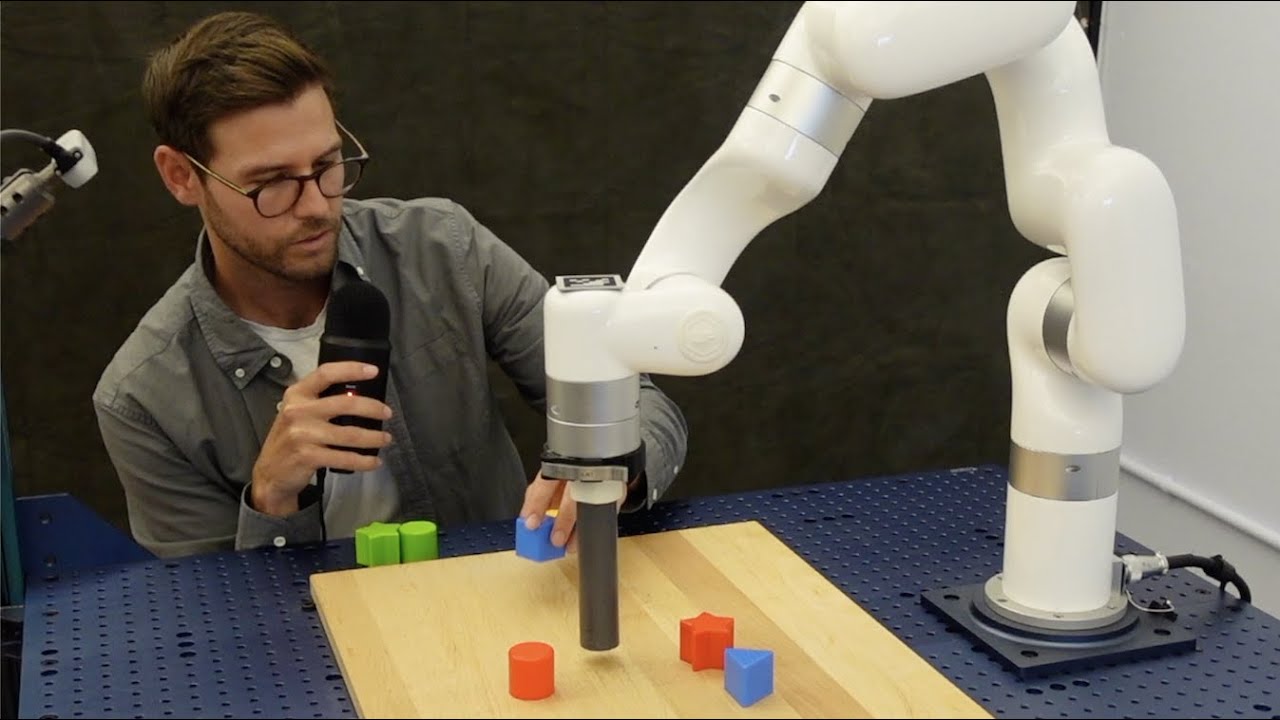 A person guiding a robot arm as it rearranges colored blocks