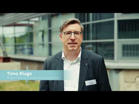Ein Mann mit kurz gescheitelten Haaren und Brille steht im Freien vor einem modernen Gebäude mit Glasfassade. Er trägt ein dunkelblaues Sakko über hellem Hemd und blickt direkt in die Kamera. Links unten ist in einem hellblauen Balken ein eingeblendeter Name und die Funktionszeile „Mitglied des Vorstandes – Sparkasse Essen“ zu sehen, wie in einem Videostandbild. Die Stimmung ist sachlich, leicht bewölktes Tageslicht.