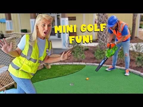 Two people on a mini golf green: a surprised woman in a yellow safety vest watches as a worker in a blue helmet uses a tool toward a small orange ball, with 'MINI GOLF FUN!' text overlay.
