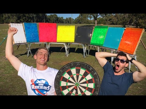Two men pose outdoors with a row of colorful boards and a large dartboard between them, smiling.",