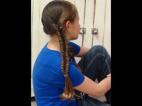 Girl with two clean fishtail braids, showcasing the Glimmer Fish Braid hairstyle, seated against a backdrop of wooden panels, wearing a blue shirt and jeans.