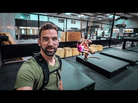 Man wearing a harness takes a selfie in an indoor obstacle-course gym; two kids swing on ropes in the background.