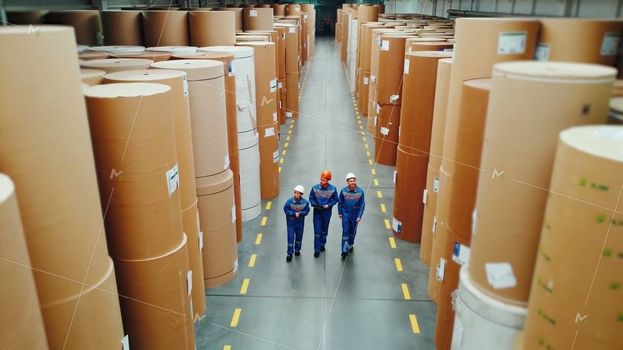 Paper mill workers walk through a huge warehouse with large rolls of paper. Engineers are taking