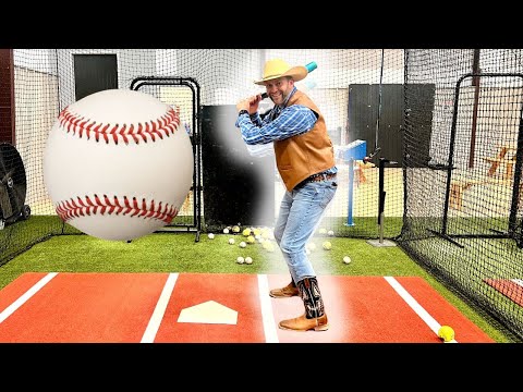 Man in a cowboy hat and vest batting in a indoor cage, swinging at a giant oversized baseball balloon.