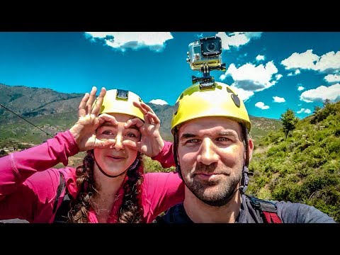 Two hikers wearing yellow helmets take a selfie outdoors, with a GoPro mounted on one helmet and mountain scenery behind them.
