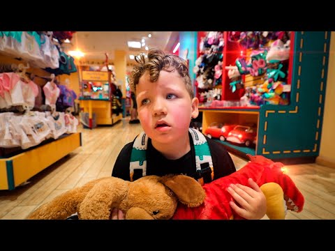 Young boy in a toy store holding a brown teddy bear and a red plush dragon, looking away with a backpack on.