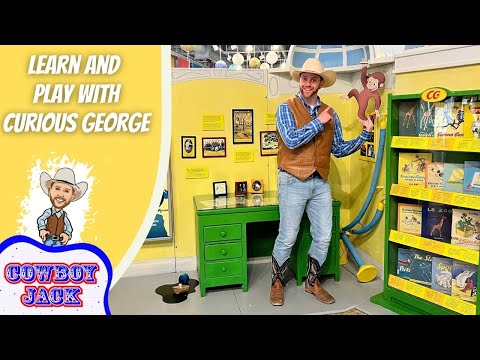 Man in a cowboy hat and vest poses beside a Curious George exhibit with a green desk and yellow wall.
