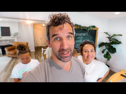 Man in the foreground takes a selfie with two smiling people in a bright, cozy living space behind him