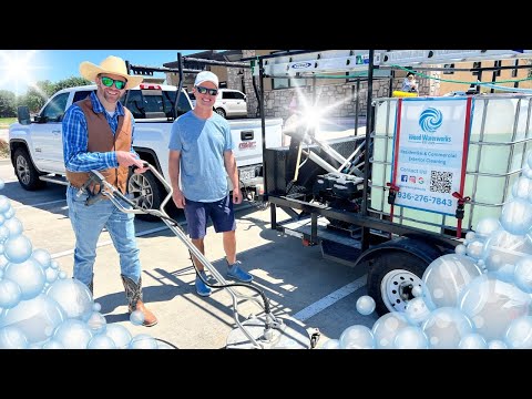 Two workers in hats pose beside a trailer-mounted pressure washer advertising exterior cleaning services in a parking lot with a white pickup nearby.