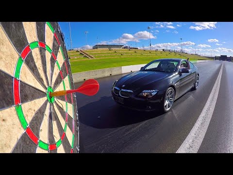 Black BMW convertible on a drag strip with a large dartboard in the foreground, a red dart near the bullseye as the car approaches.