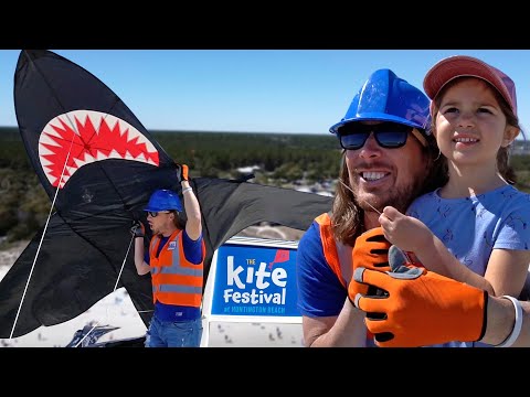Man wearing a blue hard hat and orange vest with a young girl on the beach, as a large black shark-shaped kite unfolds for Huntington Beach's Kite Festival in the background.