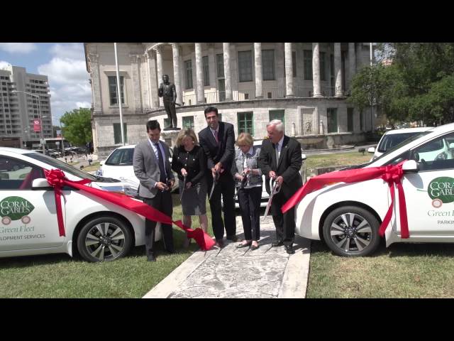 City leaders during a ribbon cutting ceremony. The ribbon is strung between two electric vehicles. 