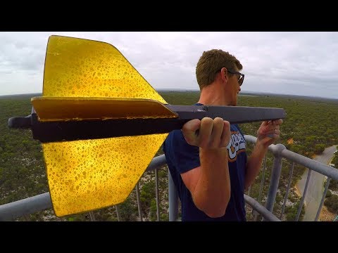 Man wearing sunglasses on a railing holds a large yellow arrow-shaped object toward the camera, overlooking a forested landscape.