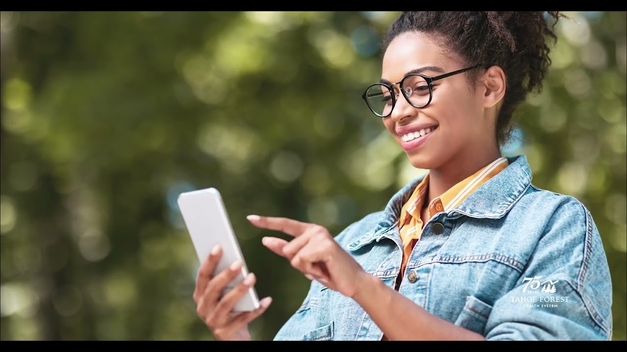 Woman wearing glasses looking at a phone with blurred trees in the background