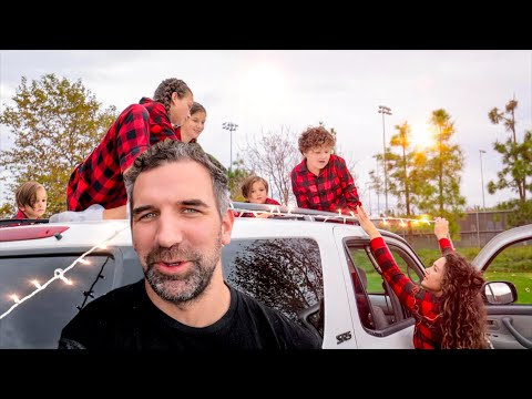 Family with plaid shirts decorating a white SUV with string lights in a park setting at sunset.