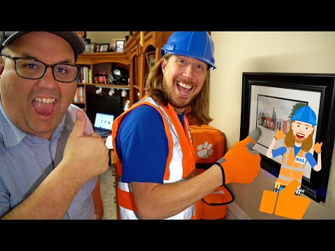 Two men smiling and giving thumbs up; one in a blue hard hat and orange safety vest, the other in a cap, in a home workshop backdrop with a framed painting and paw-print bag.