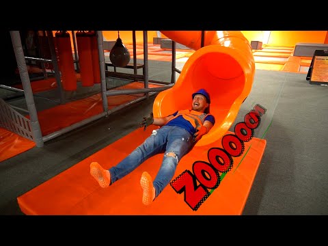 Man in a blue helmet and orange safety vest slides down a bright orange indoor tube slide, arms raised in excitement at a trampoline park, with a padded orange mat beneath.
