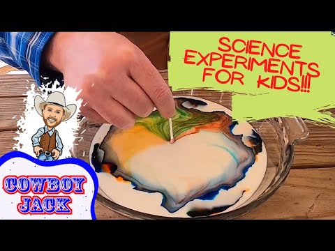 Hand swirls neon colors in a shallow dish for a kids' science experiment. A toothpick guides the mixing on a wooden table.