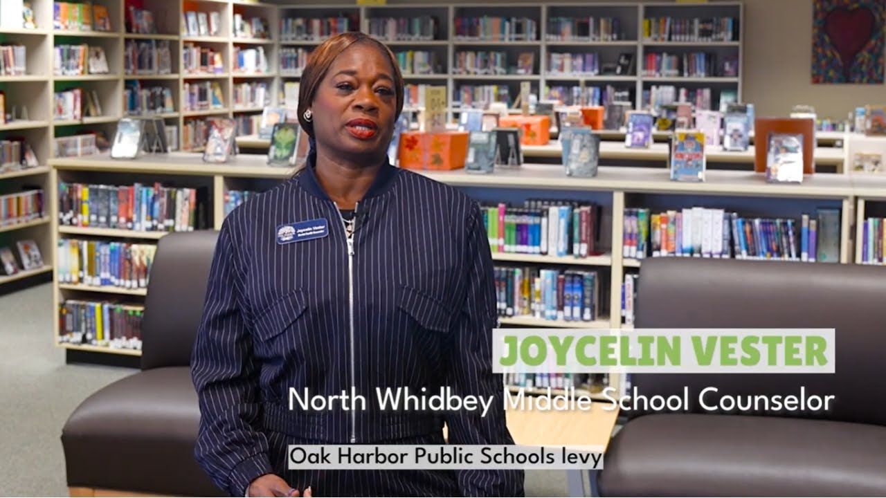 Video preview image of mental health counselor Joycelin Vester in a blue pinstriped jacket stands in a school library; click to visit the ParentSquare post with the video