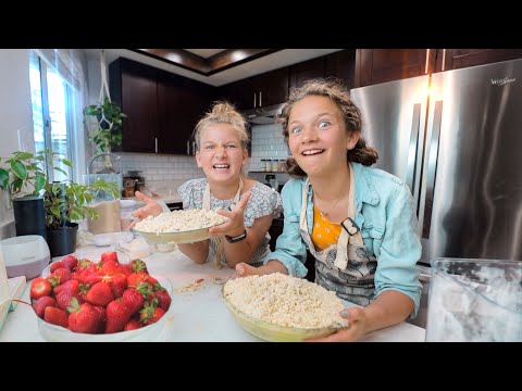 Two girls bake together in a bright kitchen, smiling while holding bowls of crumb-topped pie with fresh strawberries on the counter nearby.