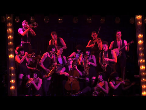Spotlit cabaret chair routine—gloves, top hat, and a smirk: the revival’s sound in one image