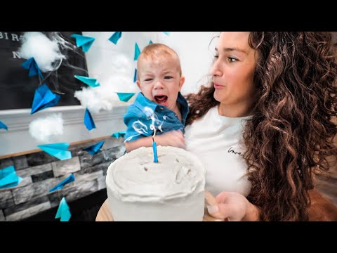 Mom holds a crying baby beside a white frosted cake with a single blue candle during a birthday celebration.