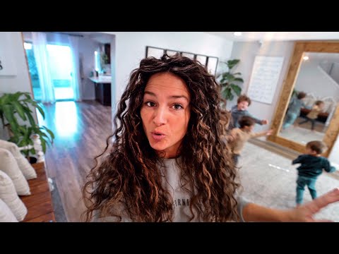 Woman with long curly hair takes a selfie in a bright home, while children play in the background.