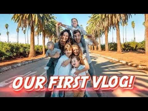 Family of six posing on a sunny palm-lined street with a bold 'OUR FIRST VLOG!' banner across the bottom.