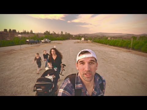 Man in a white cap takes a selfie at the front while a family with a baby stroller walks along a dusty road at sunset.