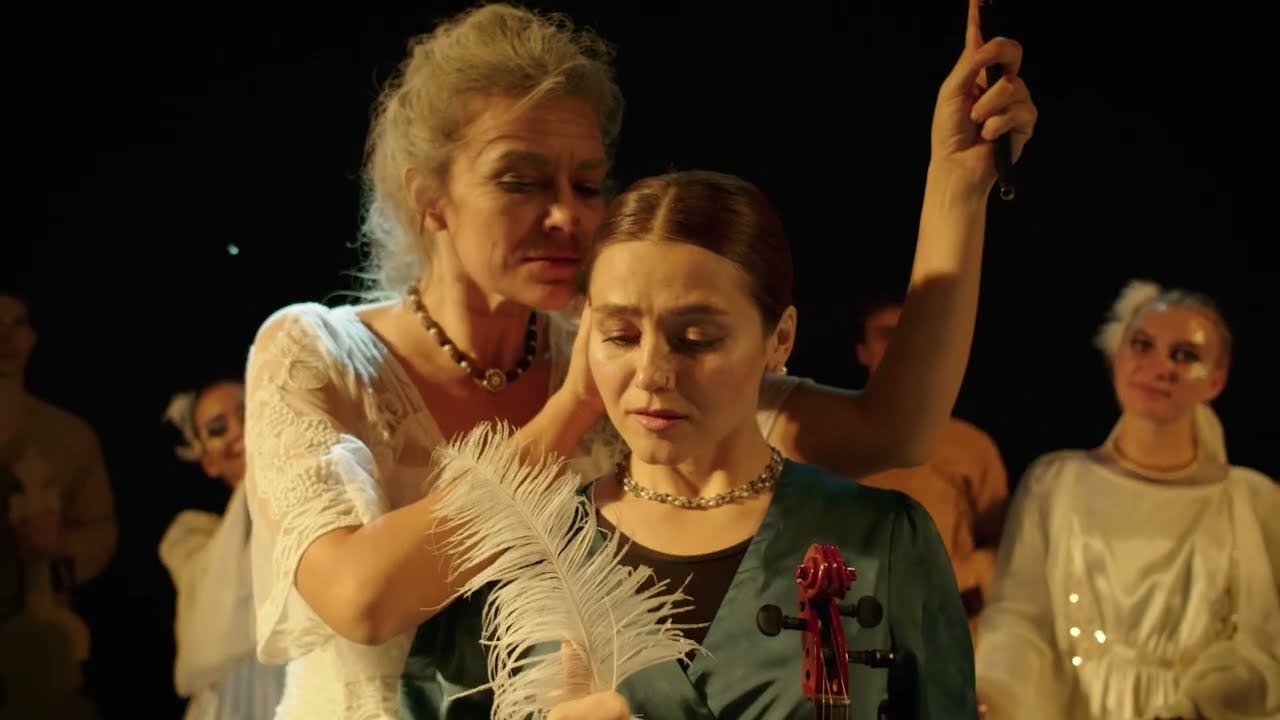 Two performers on a darkened stage, lit warm against black: an older woman stands behind a younger one, who holds a violin and a long white feather.