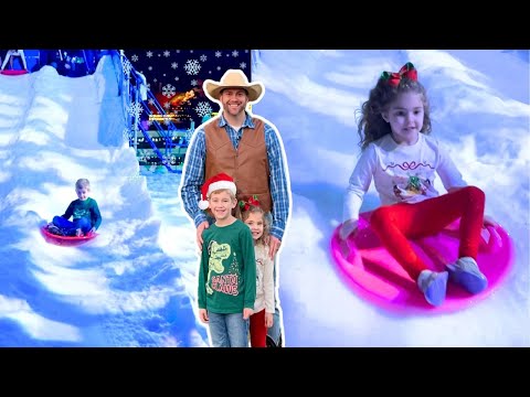 Smiling man in a cowboy hat and vest poses with two children at a snowy indoor slide, while others sled nearby under snowflakes.