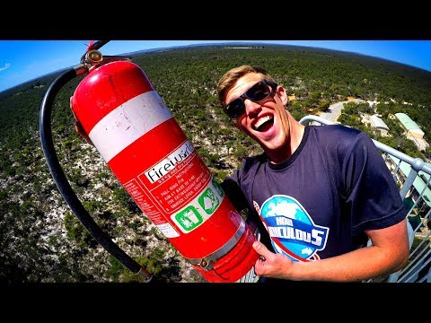Man wearing sunglasses grins at the camera while holding a large red fire extinguisher outdoors on a viewing platform.