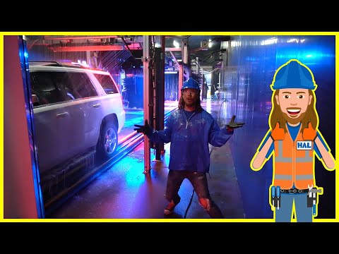 Person in blue rain gear and hard hat standing in a wet industrial hallway with a white SUV on a track nearby; colorful lights around.